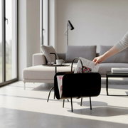 Schwarzer Metall-Zeitungsständer mit Griff und vier Beinen, gefüllt mit Magazinen, im hellen Wohnzimmer neben Sofa und Beistelltisch, perspektivisch in leichter Seitenansicht von vorn