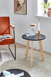 Runder Beistelltisch mit schwarzer Tischplatte und vier hellen Holzbeinen, seitliche Perspektive im Wohnraum neben einem orangefarbenen Sessel