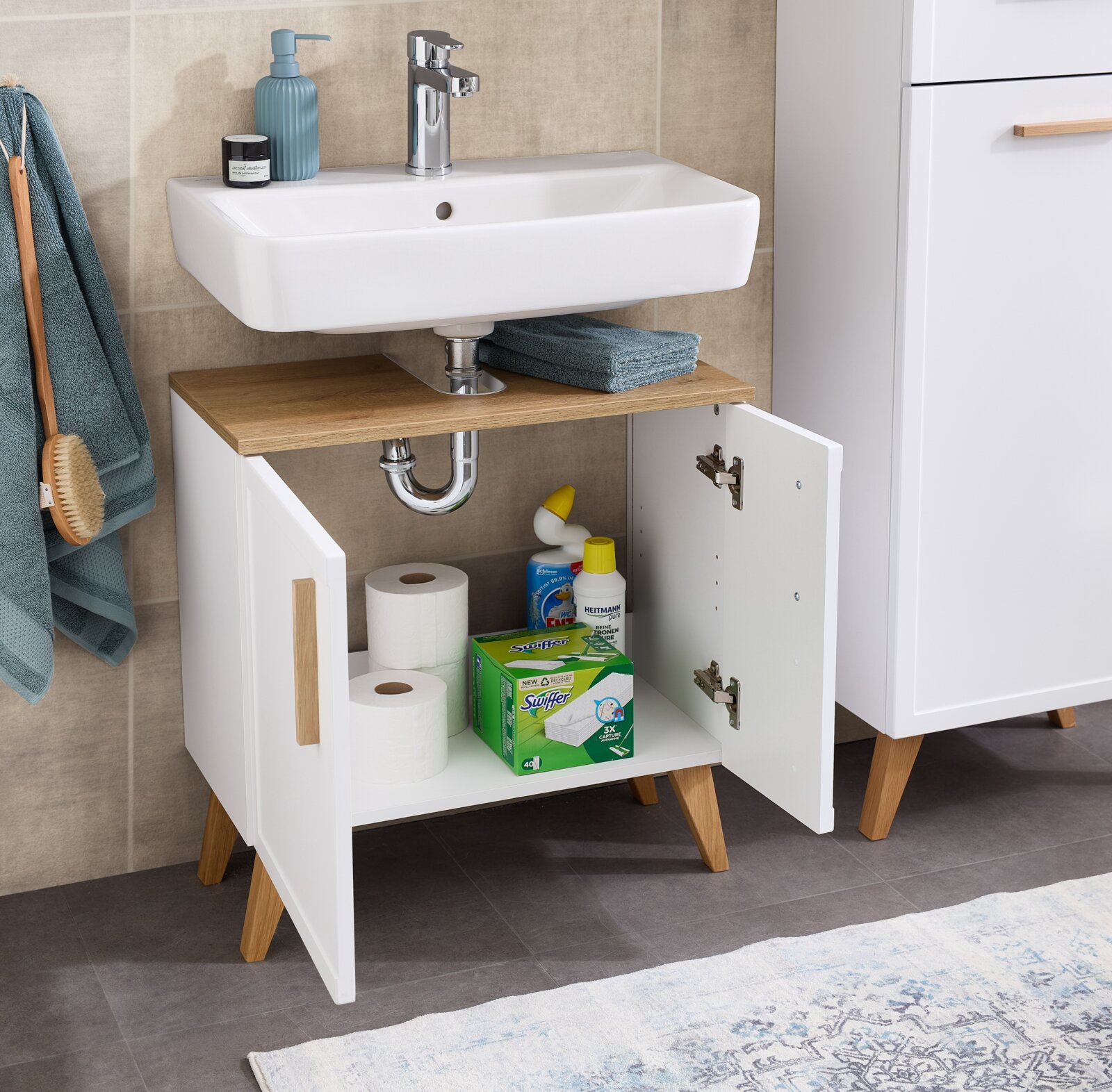 Waschbeckenunterschrank mit offenen Türen, gefüllt mit Reinigungsmitteln und Toilettenpapier, aus einer frontalen Perspektive fotografiert.