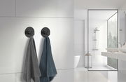 Zwei schwarze Edelstahlhaken an einer weißen Badezimmerwand, von denen jeweils ein Handtuch hängt. Die Perspektive zeigt die Haken von vorne in einem modernen Badezimmer.