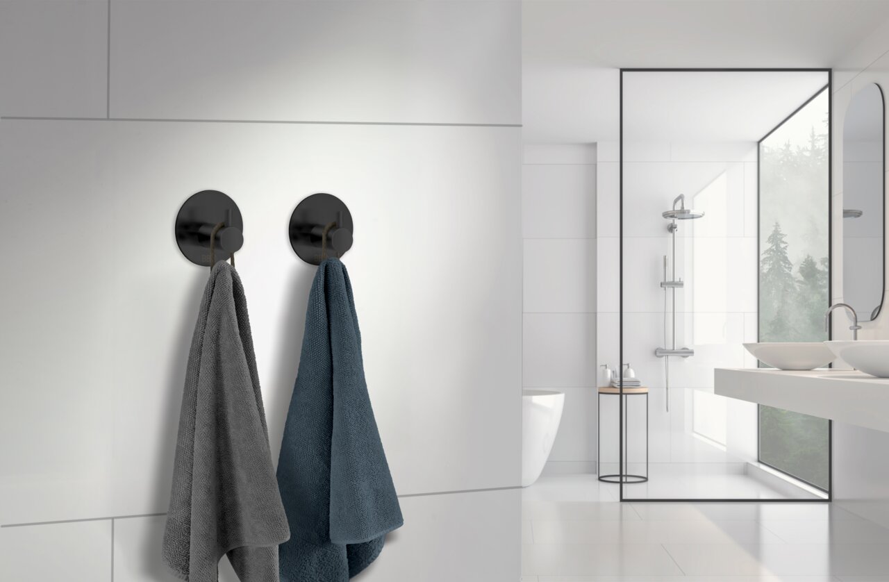 Zwei schwarze Edelstahlhaken an einer weißen Badezimmerwand, von denen jeweils ein Handtuch hängt. Die Perspektive zeigt die Haken von vorne in einem modernen Badezimmer.