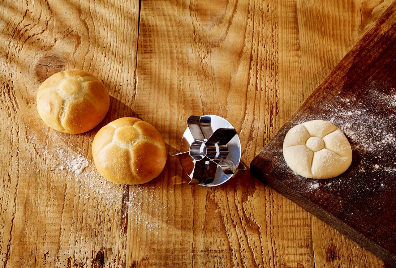 Een metalen broodstempel op een houten tafel, omringd door gevormde en ongevormde deegballen, gezien vanuit vogelperspectief.