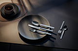 Elegantes, mattes Besteckset auf einem dunklen Tisch, bestehend aus Messer, Gabel und Löffel, stilvoll arrangiert auf einem Teller. Perspektive von oben.