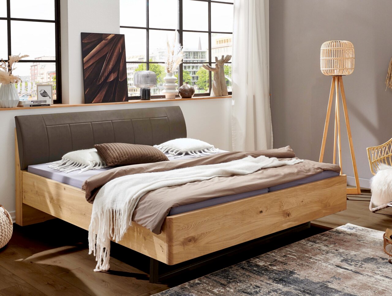 Linea Natura Comfortabel bed CARDIFF Modern houten bed met een grijs gestoffeerd hoofdeinde en een frame van naturel massief hout, bekleed met beige beddengoed en een deken, in een lichte slaapkamer met grote ramen; perspectief: schuin van voren (3/4-aanzicht)