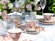 Gedeckter Tisch im Freien mit rot-weiß gemusterten Porzellan-Teetassen und Untertassen, Wassergläsern, goldenen Löffeln und Blumenvasen; Perspektive: leicht erhöhte, schräge Nahaufnahme von der Seite.