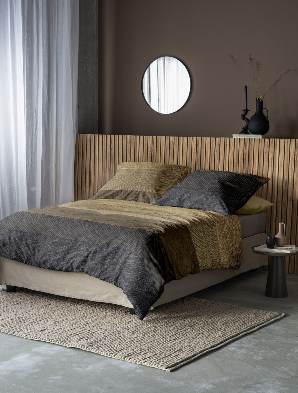 Bett mit gestreifter Bettwäsche in Erdtönen, seitliche Perspektive, modernes Schlafzimmerdesign mit Holzkopfteil und runder Wandspiegel.