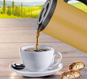 Isolierkanne in Seitenansicht, die Kaffee in eine weiße Tasse auf einem Holztisch mit Gebäck im Vordergrund und einer grünen Landschaft im Hintergrund gießt.