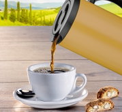 Isolierkanne in Seitenansicht, die Kaffee in eine weiße Tasse auf einem Holztisch mit Gebäck im Vordergrund und einer grünen Landschaft im Hintergrund gießt.