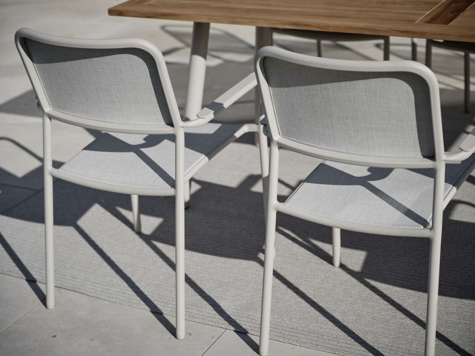 Zwei graue Stapelsessel aus Metall und Stoff, von hinten gesehen, neben einem Holztisch auf einer Terrasse.