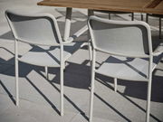 Zwei graue Stapelsessel aus Metall und Stoff, von hinten gesehen, neben einem Holztisch auf einer Terrasse.