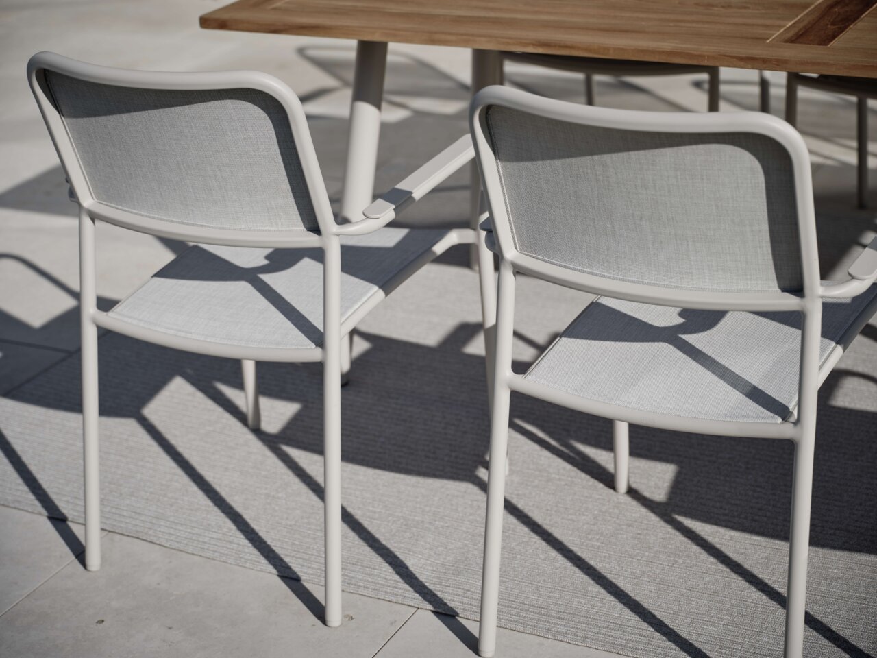 Twee grijze stapelstoelen van metaal en stof, gezien vanaf de achterkant, naast een houten tafel op een terras.