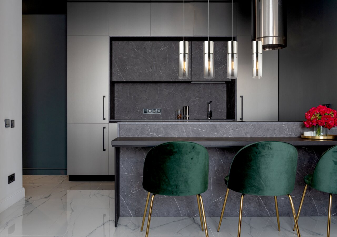 Moderne Küche mit drei eleganten Hängeleuchten über einer Theke, grüne Samtstühle und graue Schränke, seitliche Perspektive.