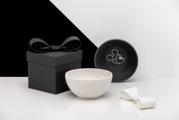 Weiße Keramikschale vor schwarzer Geschenkbox mit Schleife und runder schwarzer Dose mit Mickey-Mouse-Motiv, Studioaufnahme in frontaler Perspektive