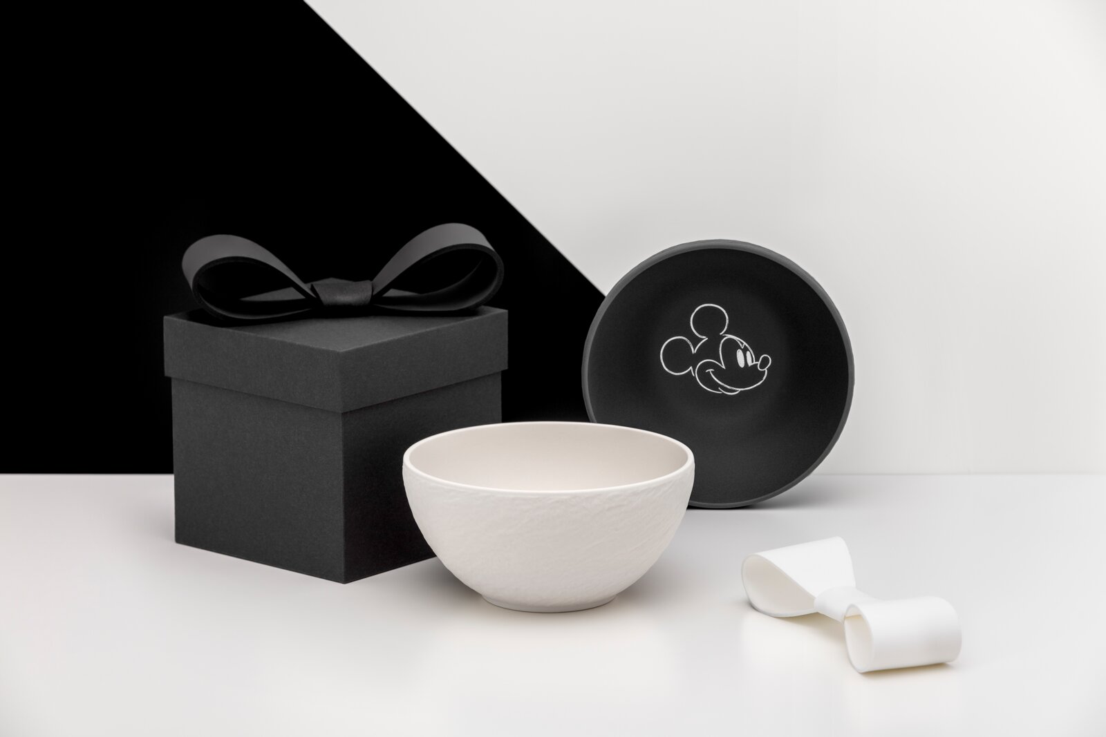 Weiße Keramikschale vor schwarzer Geschenkbox mit Schleife und runder schwarzer Dose mit Mickey-Mouse-Motiv, Studioaufnahme in frontaler Perspektive
