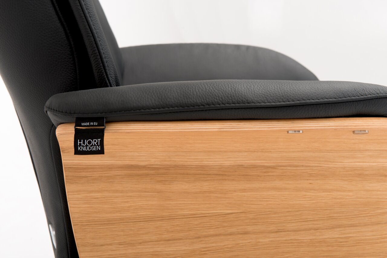 Zijaanzicht van een relaxfauteuil met zwarte lederen bekleding en houten panelen, met zichtbaar het label 'Hjort Knudsen'.