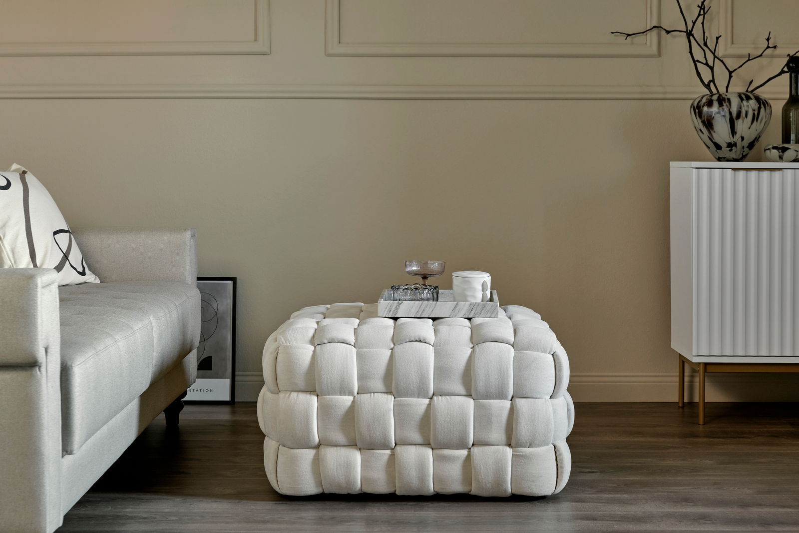 Weißer, gepolsterter Hocker in einem modernen Wohnzimmer, frontal fotografiert, neben einem Sofa und einer Kommode.