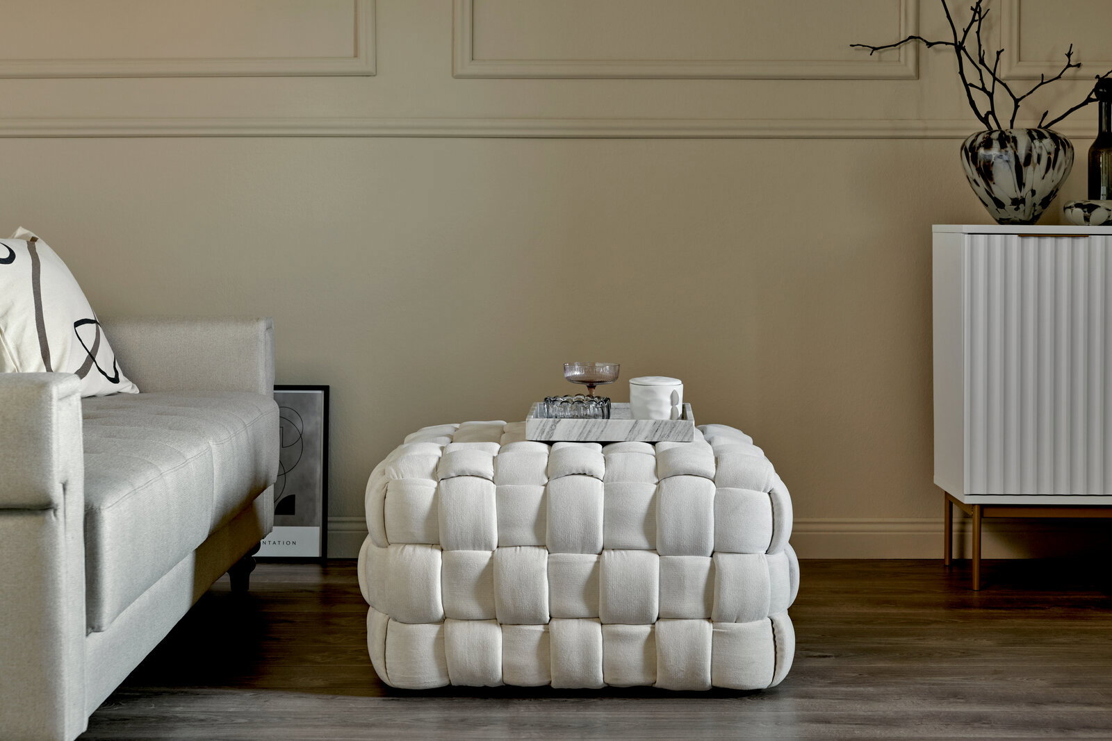 Jahnke Hocker WEB ART Weißer, gepolsterter Hocker in einem modernen Wohnzimmer, frontal fotografiert, neben einem Sofa und einer Kommode.