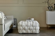 Witte, gestoffeerde poef in een moderne woonkamer, van voren gefotografeerd, naast een bank en een dressoir.