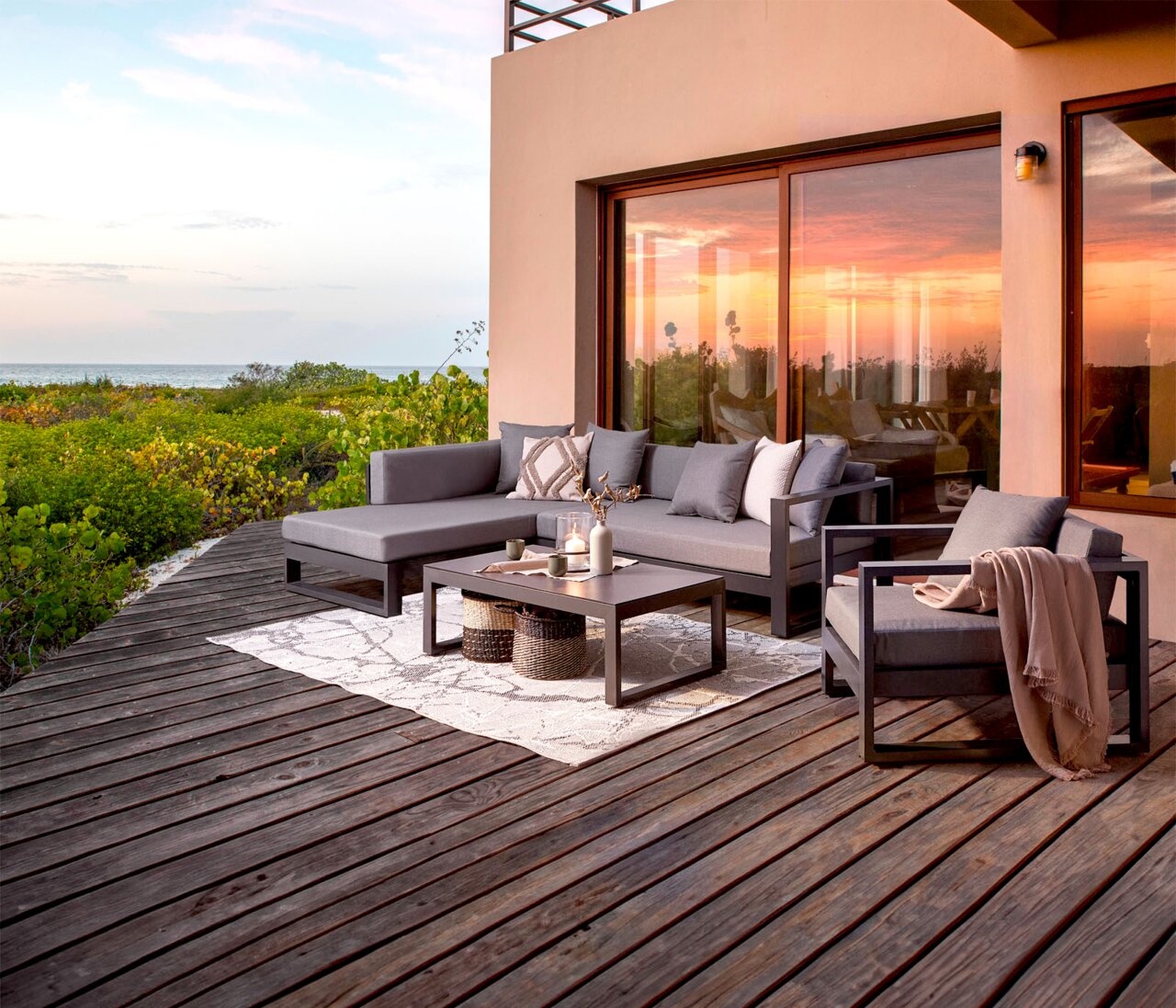 Moderne Outdoor-Lounge auf Holzterrasse mit grauem Ecksofa, Sessel und Couchtisch vor großer Glasschiebetür, seitliche Perspektive mit Blick auf Meer und Sonnenuntergang