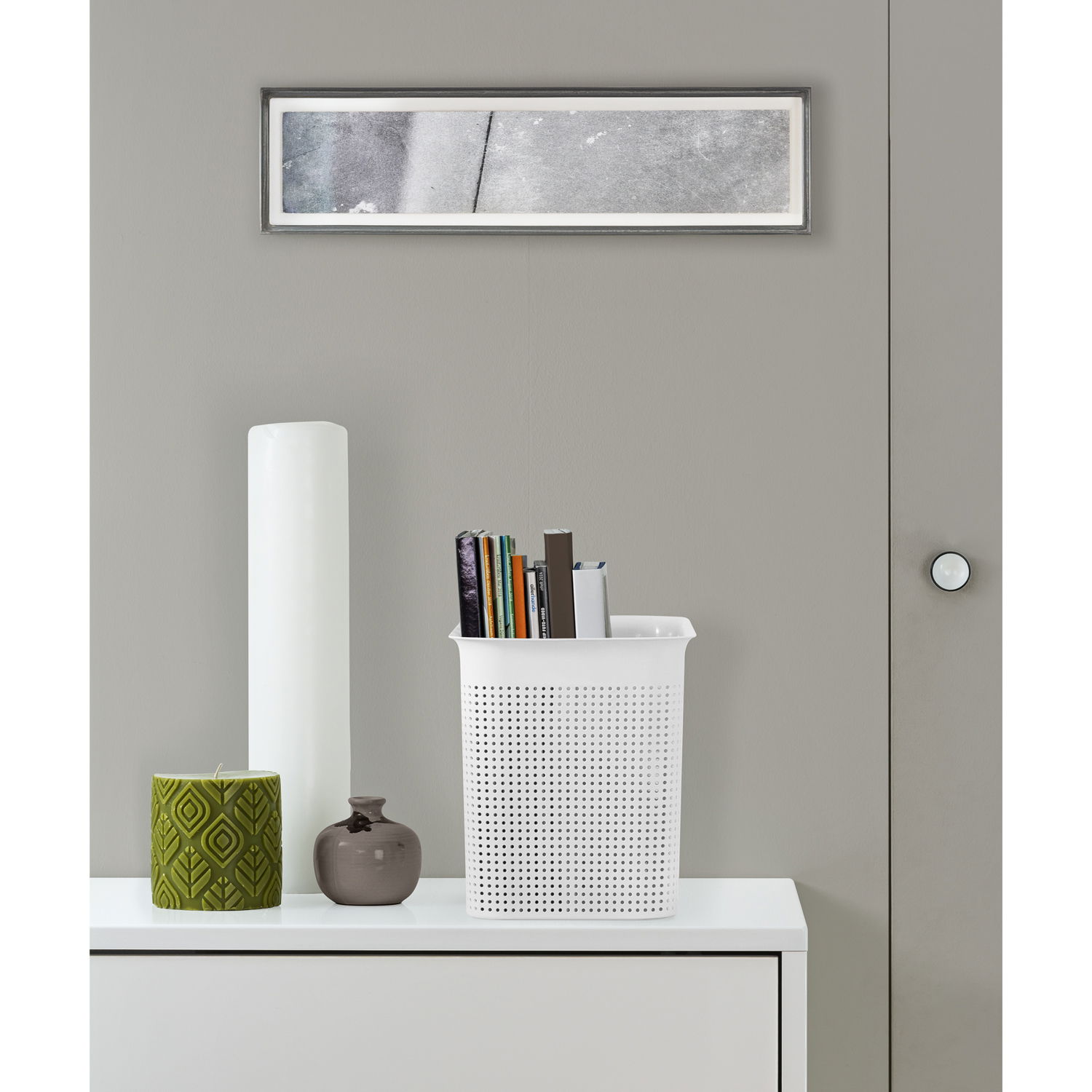 Weiße, schmale Aufbewahrungsbox mit perforiertem Design, gefüllt mit Büchern, auf einem weißen Möbelstück stehend, aus frontaler Perspektive.