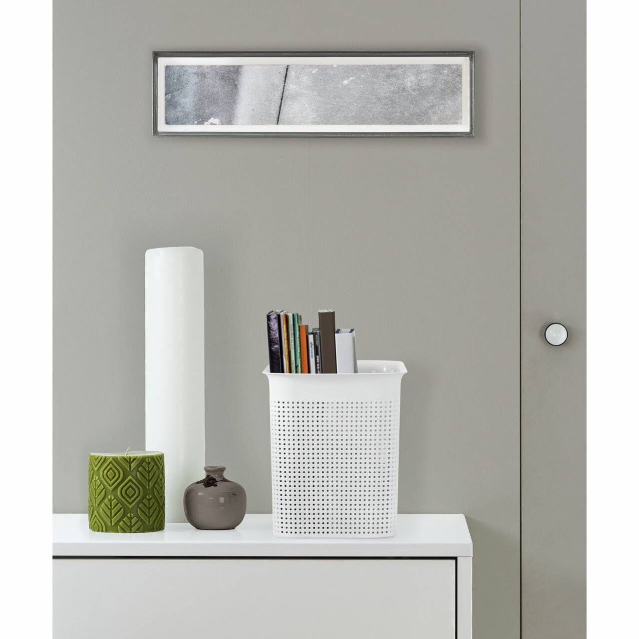 Weiße, schmale Aufbewahrungsbox mit perforiertem Design, gefüllt mit Büchern, auf einem weißen Möbelstück stehend, aus frontaler Perspektive.