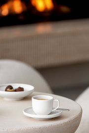 Weiße Espressountertasse mit Tasse und Löffel auf einem Tisch, im Hintergrund ein unscharfer Kamin. Perspektive von schräg oben.