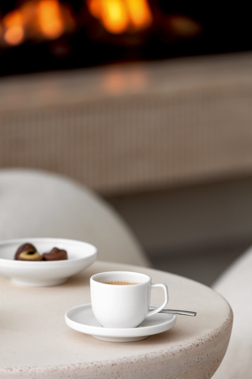 Weiße Espressountertasse mit Tasse und Löffel auf einem Tisch, im Hintergrund ein unscharfer Kamin. Perspektive von schräg oben.