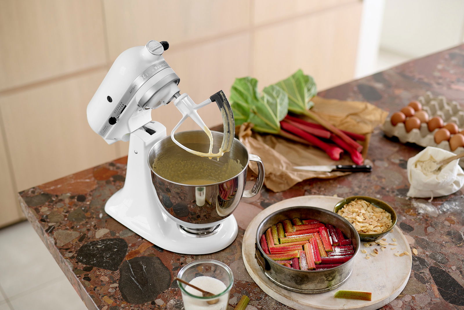 Weiße Küchenmaschine auf einer Arbeitsplatte, von der Seite fotografiert, mit Rührschüssel und Teig, umgeben von Zutaten wie Rhabarber, Eiern und Mehl.