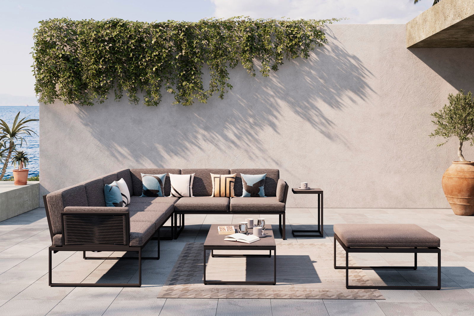 Amatio Loungegarnitur GIRONA Moderne Loungegarnitur auf einer Terrasse mit Meerblick, bestehend aus einem Ecksofa, einem Couchtisch und einem Hocker. Die Garnitur ist in einem eleganten, minimalistischen Design gehalten und von Pflanzen umgeben. Perspektive von vorne.