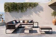 Moderne Loungegarnitur auf einer Terrasse mit Meerblick, bestehend aus einem Ecksofa, einem Couchtisch und einem Hocker. Die Garnitur ist in einem eleganten, minimalistischen Design gehalten und von Pflanzen umgeben. Perspektive von vorne.