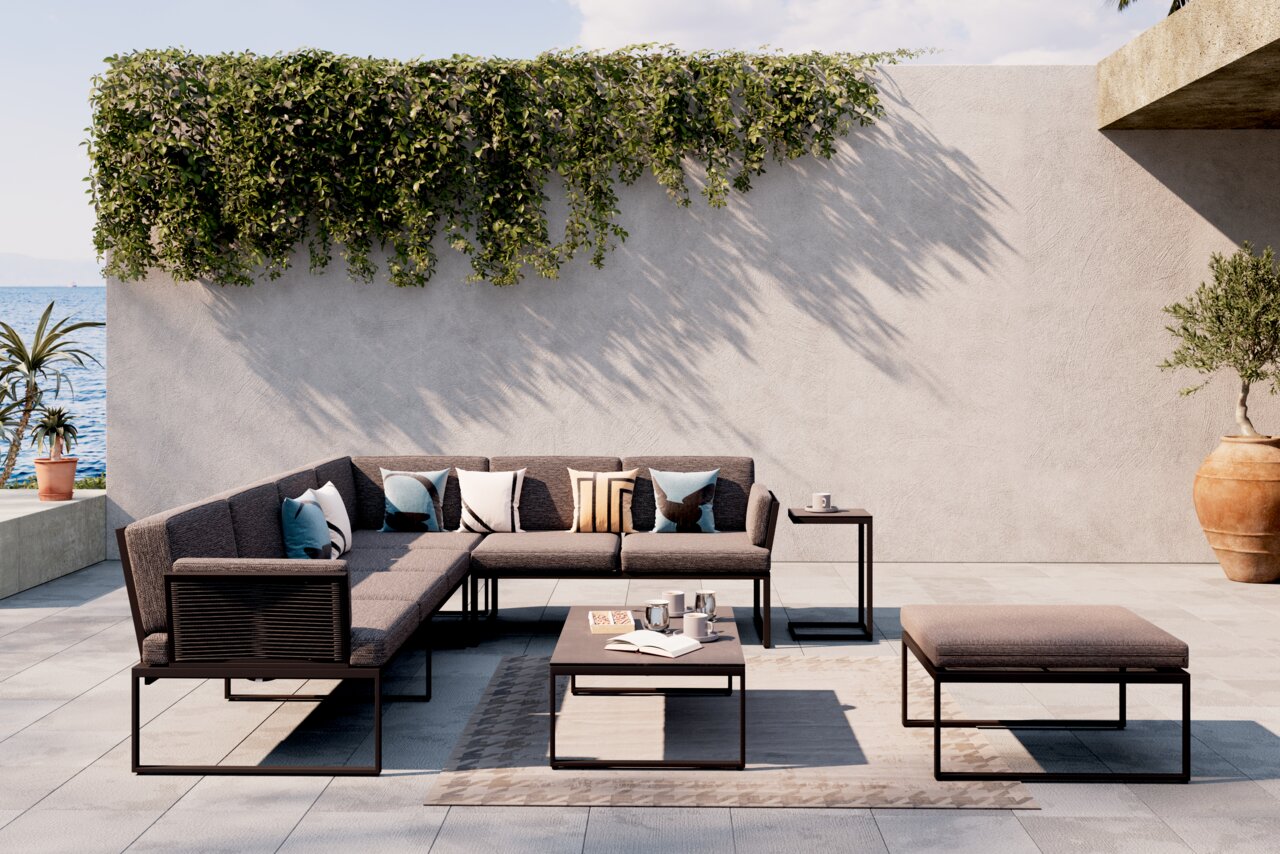 Moderne Loungegarnitur auf einer Terrasse mit Meerblick, bestehend aus einem Ecksofa, einem Couchtisch und einem Hocker. Die Garnitur ist in einem eleganten, minimalistischen Design gehalten und von Pflanzen umgeben. Perspektive von vorne.