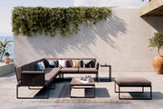 Moderne Loungegarnitur auf einer Terrasse mit Meerblick, bestehend aus einem Ecksofa, einem Couchtisch und einem Hocker. Die Garnitur ist in einem eleganten, minimalistischen Design gehalten und von Pflanzen umgeben. Perspektive von vorne.