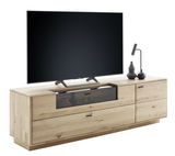 Perspektive von vorne links auf das Linea Natura Lowboard GEORGIA aus hellem Holz mit Schubladen und einem Fach mit Glastür, dekoriert mit einer Tischlampe und Vasen, darauf steht ein großer Flachbildfernseher.