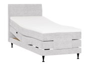 Lichtgrijs gestoffeerd elektrisch verstelbaar bed met hoog hoofdbord, wit matras, zwarte poten en open opberglade, schuin van voren weergegeven.