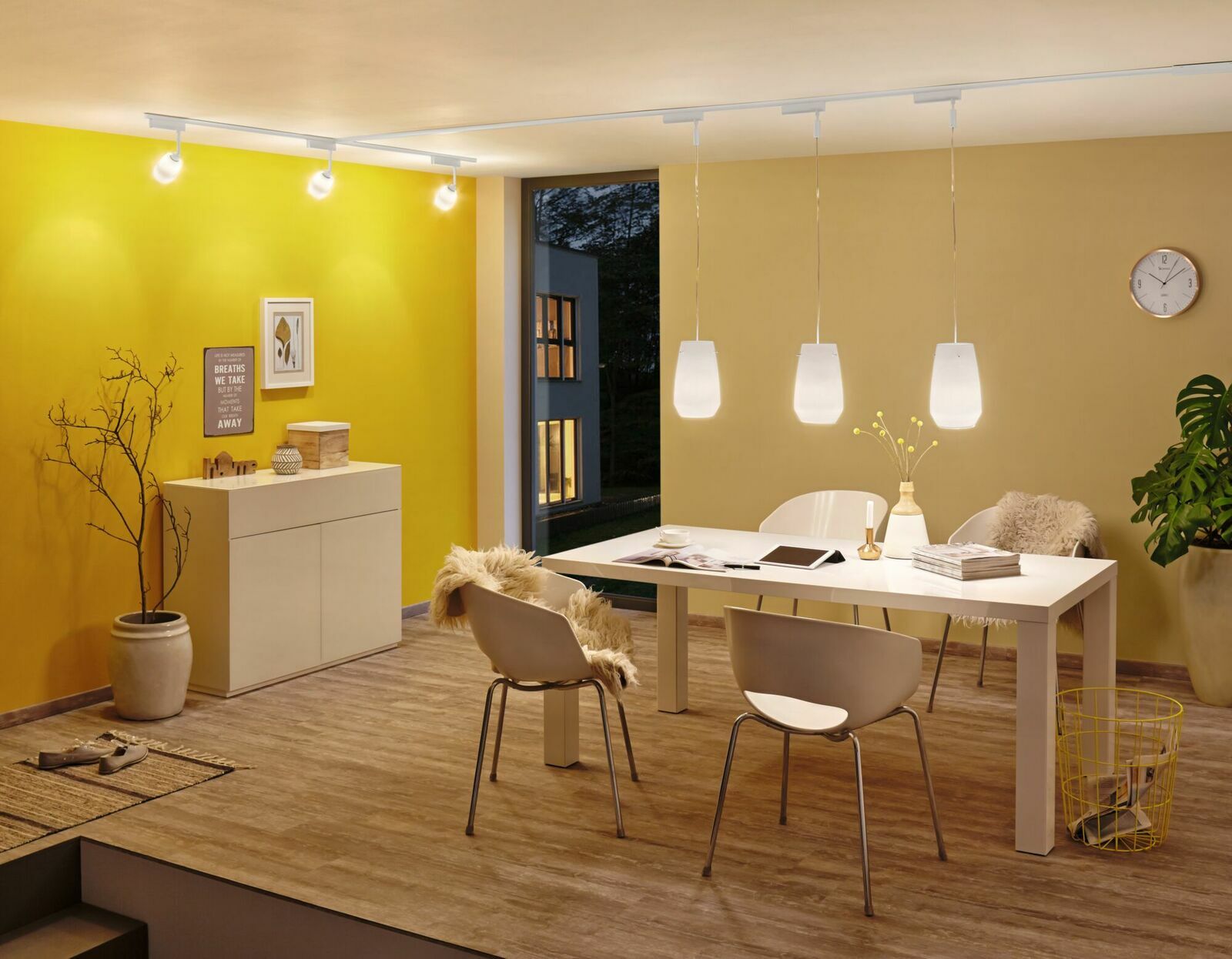 Moderne Wohnzimmerszene mit stilvoller Beleuchtung, bestehend aus einer Reihe von Deckenstrahlern und drei hängenden Lampen über einem Esstisch. Perspektive: seitliche Ansicht des Raumes.