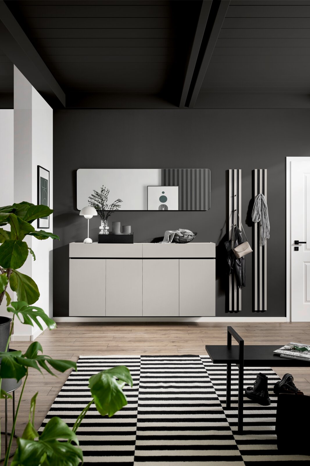 Frontalansicht einer modernen Garderobenkombination mit schwebendem Sideboard in Greige, abgerundetem Wandspiegel und schwarzen Paneel-Kleiderhaken in stilvollem Eingangsbereich.