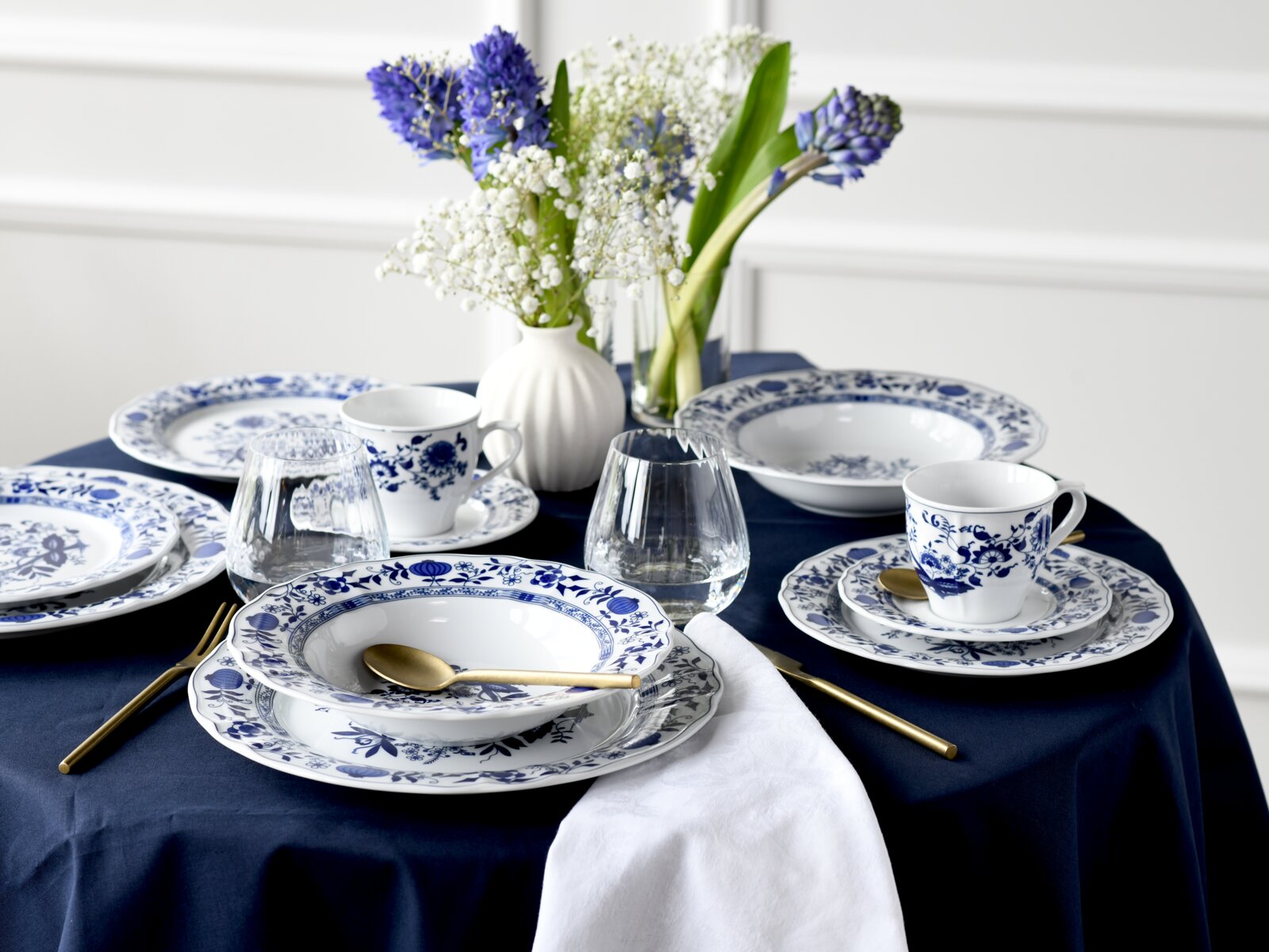 Gedeckter Tisch mit blau-weißem Porzellangeschirr im floralen Muster, goldfarbenem Besteck, Wassergläsern und Blumenvasen auf dunkelblauer Tischdecke, perspektivisch aus leicht erhöhter schräger Vorderansicht