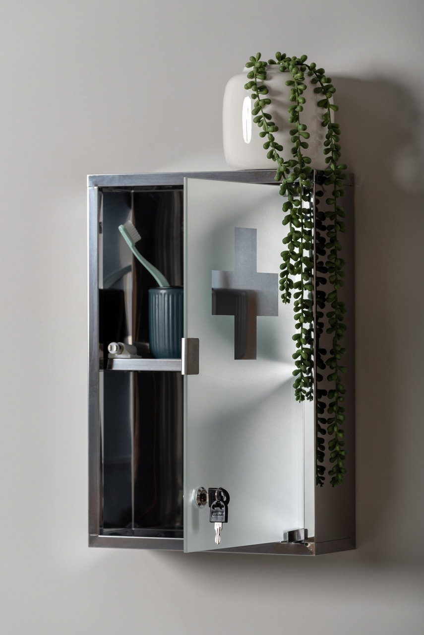 Medizinschrank aus Metall mit Glastür, teilweise geöffnet, mit einem Zahnputzbecher und Zahnbürste im Inneren, dekoriert mit einer Pflanze auf der Oberseite. Perspektive von vorne.