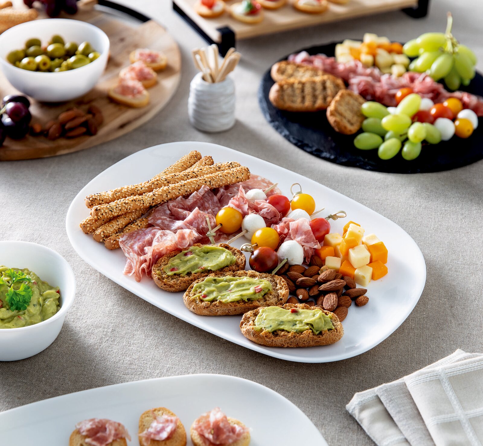Schräg von oben fotografierte Antipasti-Platte auf weißer Servierplatte mit Salami und Schinken, Sesam-Grissini, Käsewürfeln, Mandeln, Spießen mit Tomaten und Mozzarella sowie Brotscheiben mit Avocadocreme auf gedecktem Tisch