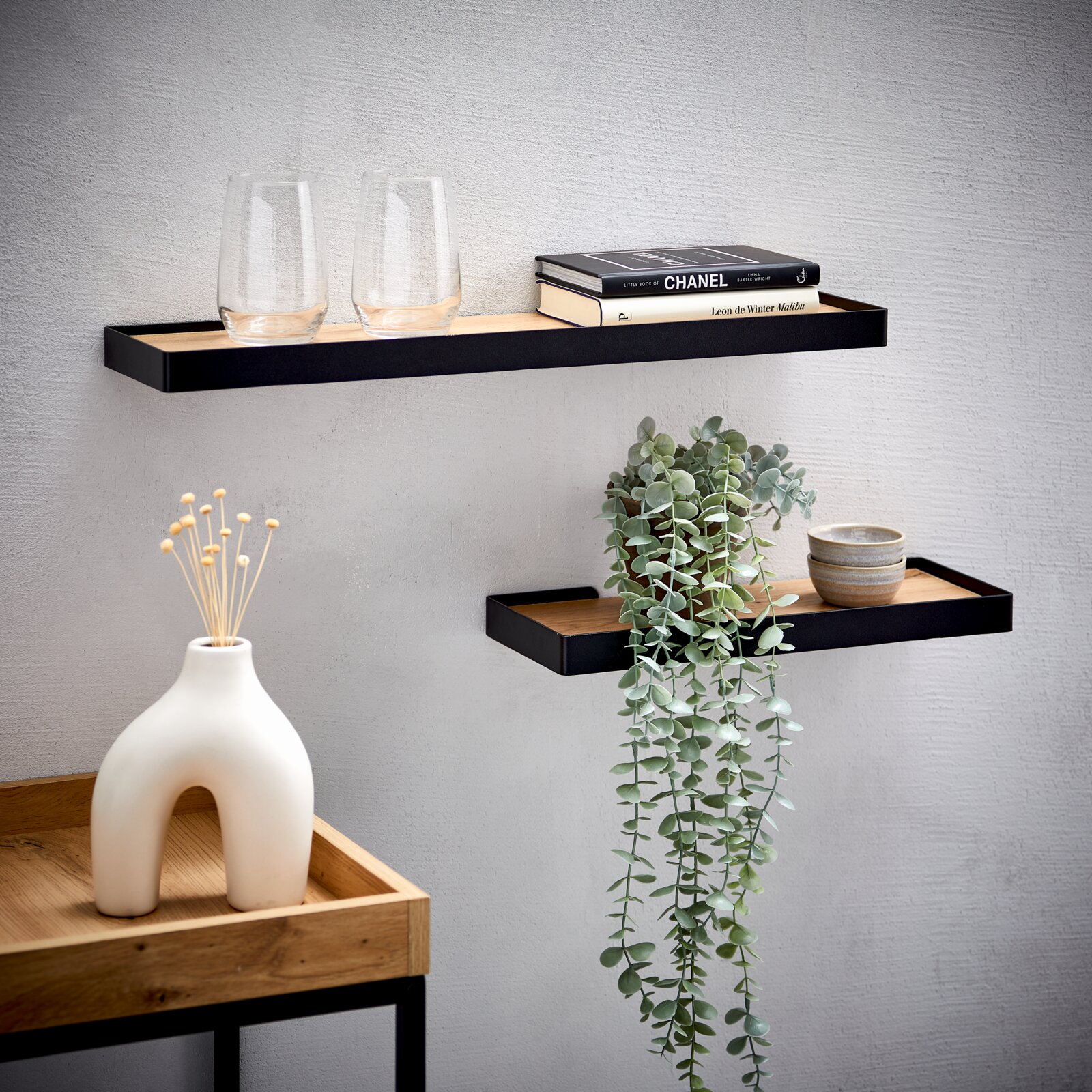 Twee moderne zwarte wandplanken met houten inleg, van voren in lichte zijaanzichtperspectief, stijlvol gedecoreerd met glazen, boeken, schaaltjes en een hangplant.