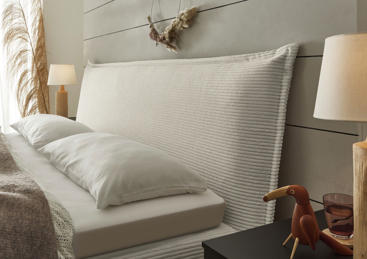 Seitliche Perspektive eines stilvollen Boxspringbetts mit geripptem Kopfteil, dekoriert mit Kissen und umgeben von eleganten Nachttischlampen und Dekorationen.