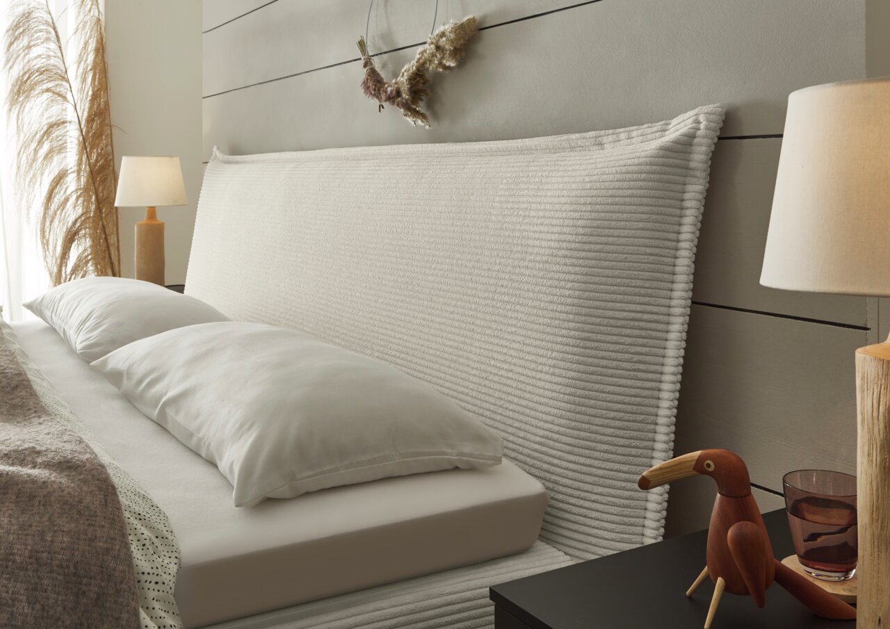 Seitliche Perspektive eines stilvollen Boxspringbetts mit geripptem Kopfteil, dekoriert mit Kissen und umgeben von eleganten Nachttischlampen und Dekorationen.