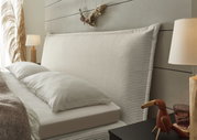 Seitliche Perspektive eines stilvollen Boxspringbetts mit geripptem Kopfteil, dekoriert mit Kissen und umgeben von eleganten Nachttischlampen und Dekorationen.
