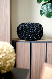 Schwarze Vase mit blütenartiger Struktur, seitliche Perspektive, auf einem Holzregal platziert.