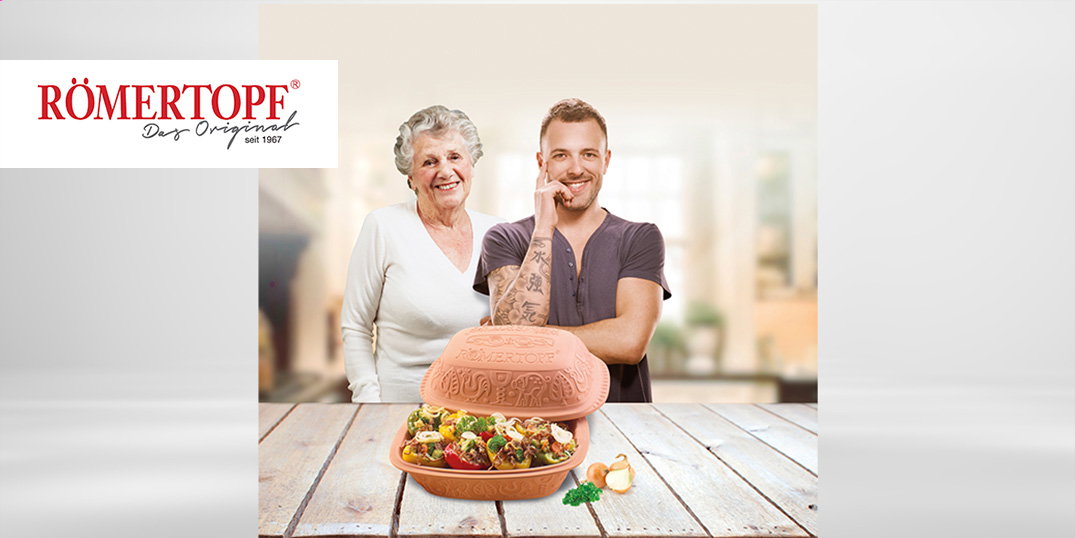 Ein Mann und eine ältere Frau stehen hinter einem Römertopf mit Essen auf einem Holztisch.