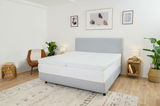 Schlafzimmer mit einem grauen Boxspringbett, das mit einem Topper ausgestattet ist. Der Raum ist modern eingerichtet mit Pflanzen, einem Sessel und Wanddekorationen. Perspektive von der Seite des Bettes.