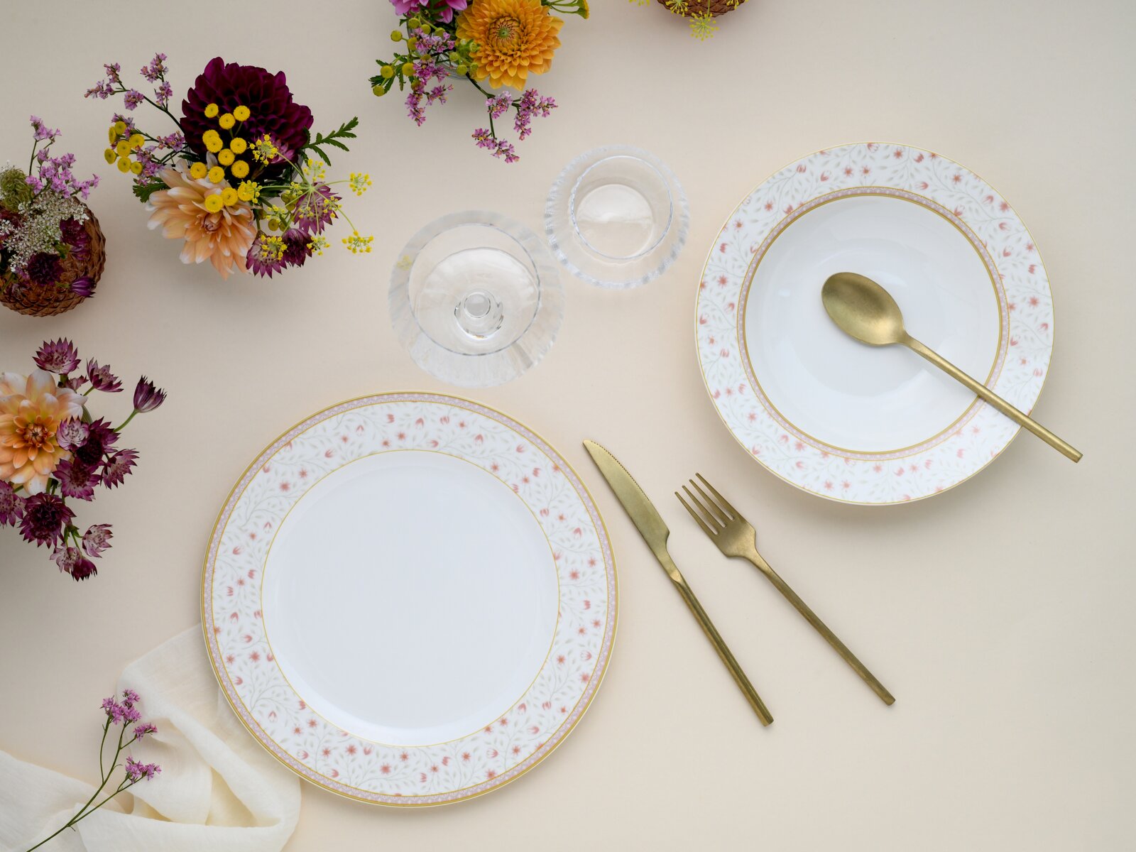 Draufsicht auf elegant gedeckten Tisch mit zwei weißen Porzellantellern mit zartem Blumenmuster und Goldrand, goldfarbenem Besteck, zwei klaren Gläsern und farbenfrohen Blumenarrangements auf hellem Hintergrund