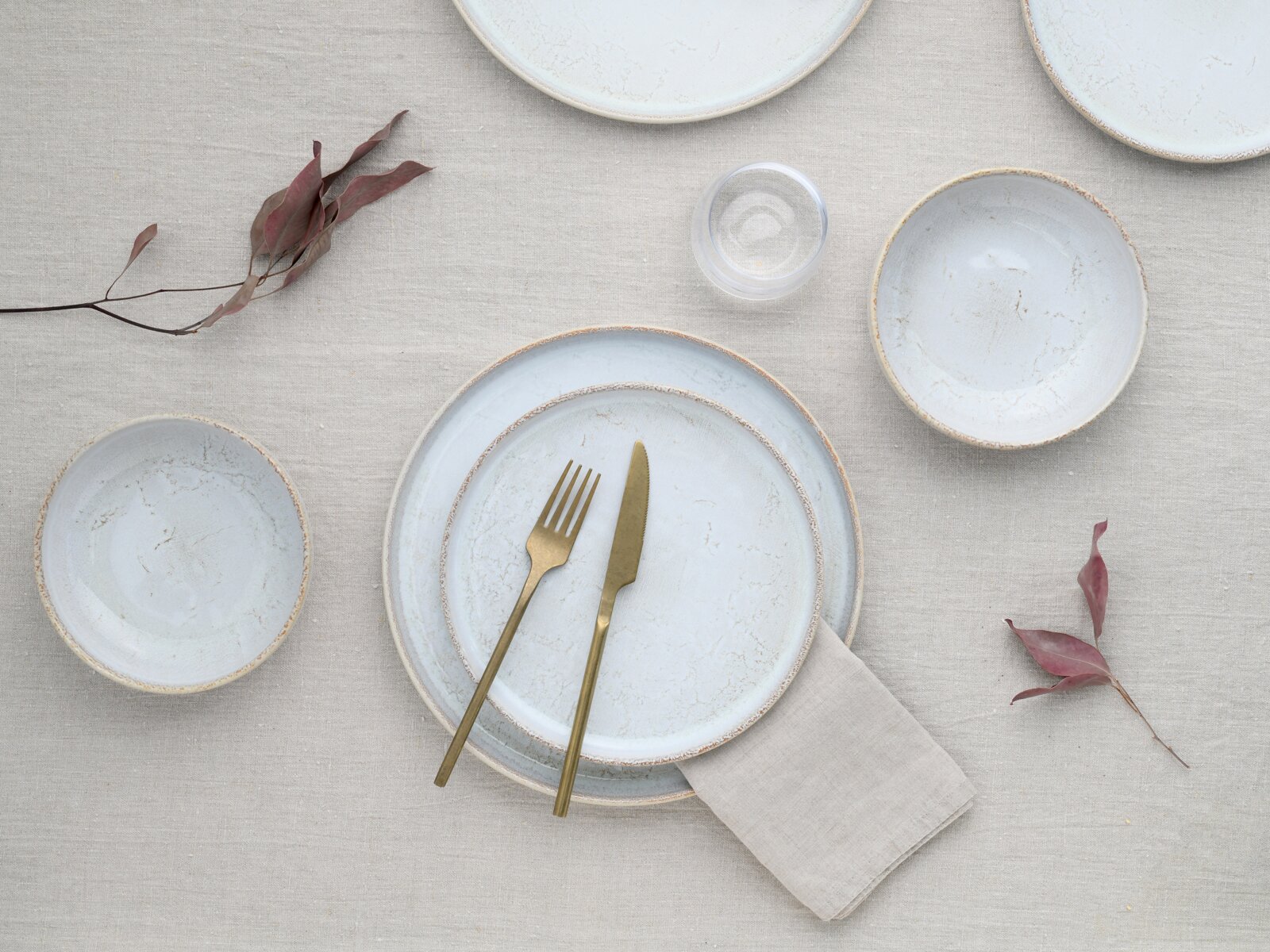 Draufsicht auf ein minimalistisches Tischgedeck mit hellblau marmorierten Keramiktellern und Schalen, goldfarbenem Besteck, Leinenserviette und Glas auf beigem Leinentuch