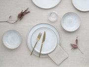 Draufsicht auf ein minimalistisches Tischgedeck mit hellblau marmorierten Keramiktellern und Schalen, goldfarbenem Besteck, Leinenserviette und Glas auf beigem Leinentuch