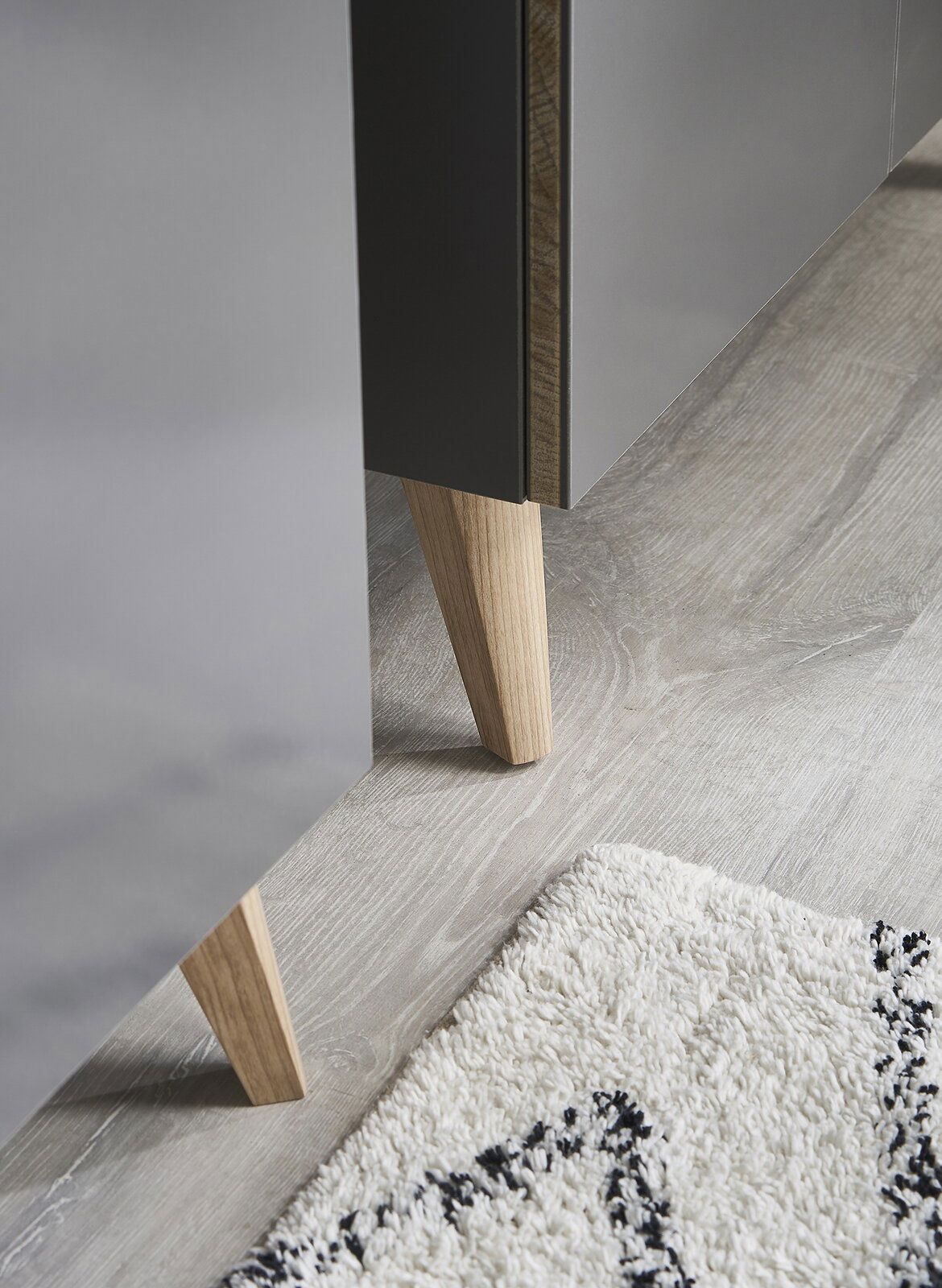Nahaufnahme eines Möbelfußes aus Holz, der an einem grauen Möbelstück befestigt ist, auf einem hellen Holzboden mit einem Teppich im Vordergrund.